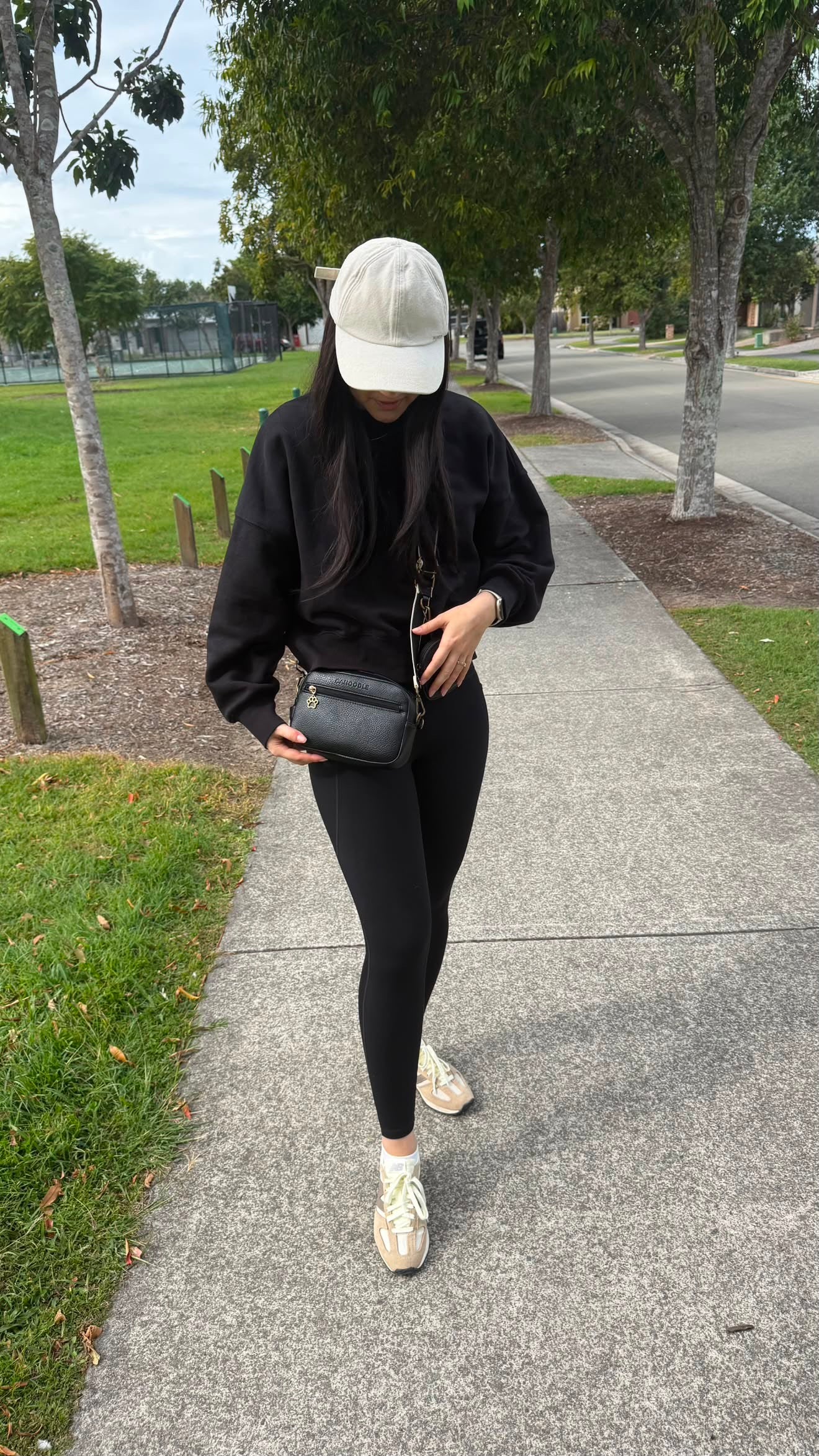 A person walking on a paved path in a park, wearing a black long-sleeve top, black leggings, a white baseball cap, and white sneakers, while carrying a small black crossbody bag.
